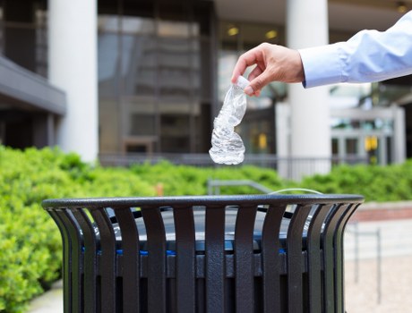 A person wearing a light blue shirt is disposing of a transparent plastic water bottle into a black outdoor rubbish bin with vertical slats, situated on a paved area beside a modern building with glass windows and white structural elements. The bin is positioned on a paved surface with visible greenery and landscaping elements nearby. The background features the urban setting of Woolwich, including parts of the building's exterior, some railings, and a walkway, indicating an outdoor public or commercial space. The scene illustrates waste disposal and rubbish collection practices relevant to local rubbish removal services offered by Office Clearance Woolwich, with the focus on responsible waste segregation and cleanliness.