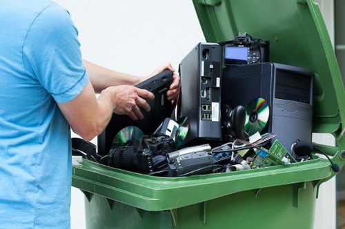 A person wearing a light blue T-shirt is seen emptying electronic waste from their hands into a large green plastic wheelie bin, which is positioned outdoors possibly on a driveway or pavement. Inside the bin, various discarded electronic items are visible, including a black desktop computer tower, a monitor, two compact discs, a digital camera with a strap, a compact digital camera, scattered circuitry, and other small electronic components, all appearing to be unused or obsolete. The scene captures the process of responsible electronic rubbish removal, highlighting the materials' plastic and metal finishes, with some items showing signs of minor wear or dust. The background reveals an outdoor environment typical of a residential or commercial property, with natural light illuminating the scene, emphasizing the collection of electronic waste ready for disposal or recycling through services like those offered by Office Clearance Woolwich. The image demonstrates the importance of proper electronic waste management within the context of rubbish removal solutions in Woolwich or nearby areas.