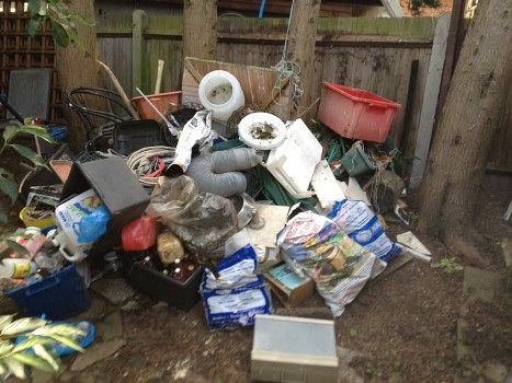 A pile of mixed household waste and rubbish situated outdoors on a patch of bare ground, consisting of various materials and objects. The collection includes plastic bags filled with waste, some crumpled and partially open, revealing discarded packaging and debris. There are several white plastic buckets, some with visible dirt or stains, stacked and leaning against other items. An orange plastic storage bin is positioned at the back of the pile, partially obscured by other objects. Also visible are black plastic garden chairs, a grey flexible drainage pipe, cardboard boxes, and a plastic container. The waste is gathered in front of a wooden fence with vertical slats, supported by wooden posts, and a few tree trunks are visible in the background. The lighting appears natural, likely from daylight, illuminating the cluttered outdoor space, which is typical of rubbish collection areas. This scene aligns with the services provided by Office Clearance Woolwich for rubbish removal within the Woolwich area, in south-east London postcode.