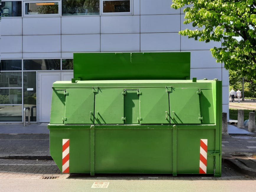 A paved outdoor area near a large industrial building features five large, rectangular, orange skip containers with green and white reflective stripes positioned along the edge of the parking space. T