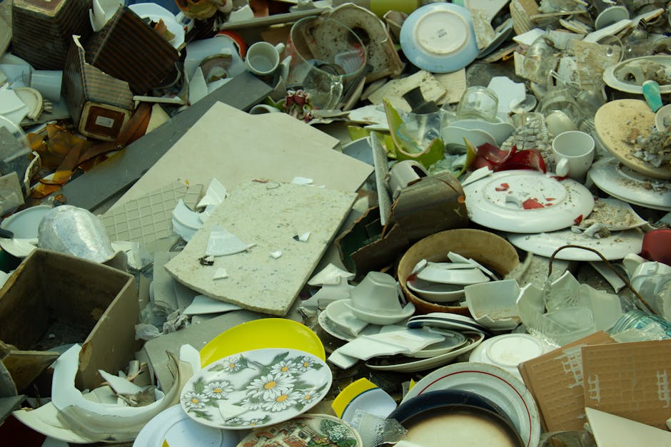A pile of black garbage bags, cardboard boxes, and discarded packaging material is stacked against a beige, textured exterior wall near a red door covered in graffiti and stickers. The bags and boxes 