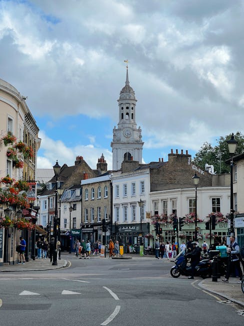 A busy town centre scene featuring a mix of historic and modern buildings, with a prominent white clock tower topped by a gold weather vane in the background. The foreground includes a paved street in