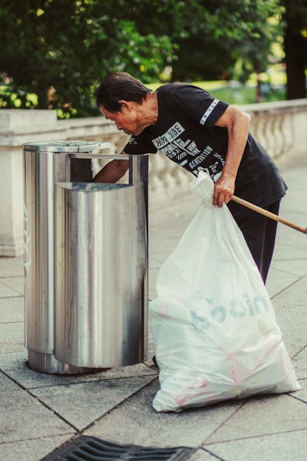 A middle-aged man with dark hair is seen outdoors, at a paved public space with a stone balustrade and green trees in the background. He is wearing a black T-shirt with white and grey printed text and