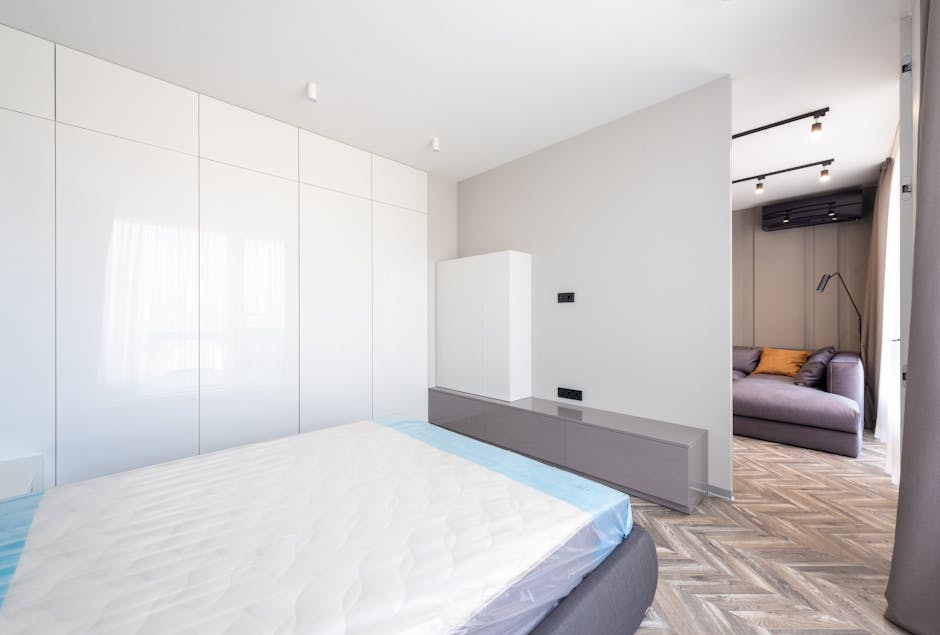 The image shows a modern, minimally decorated bedroom with a large, white wardrobe with glossy doors occupying the left side of the scene. Facing the wardrobe, part of a double bed with a plain, white